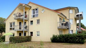 Un edificio residencial de varios pisos con balcones, techo plano y un pequeño jardín.