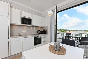 Cozinha moderna com armários brancos, micro-ondas e forno. Há uma mesa de jantar com uma cesta e uma chaleira. O quarto tem uma janela grande com vista para edifícios e árvores.