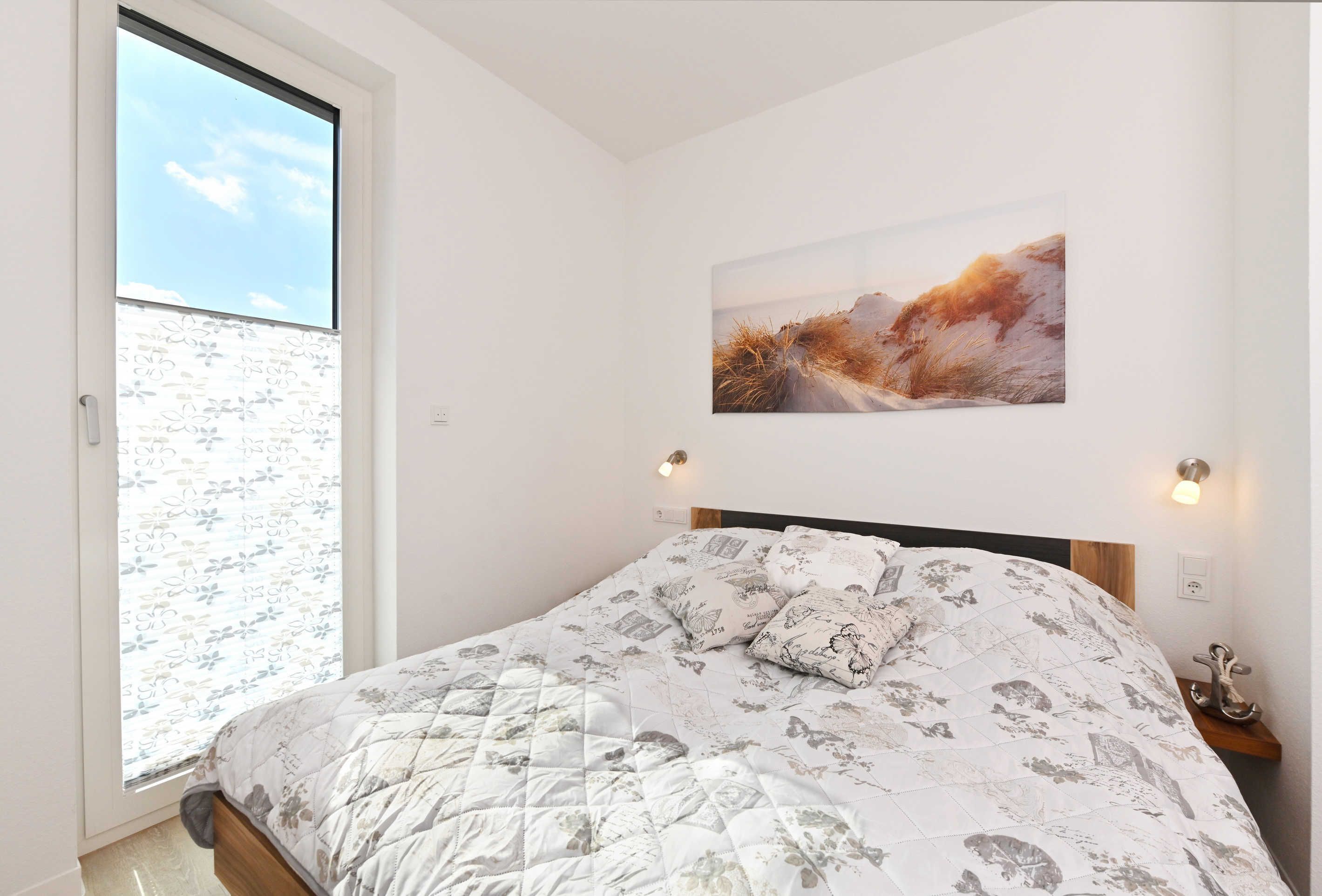A bedroom with a large window, a bed with a floral-patterned duvet, a wall-mounted painting, and bedside lamps.