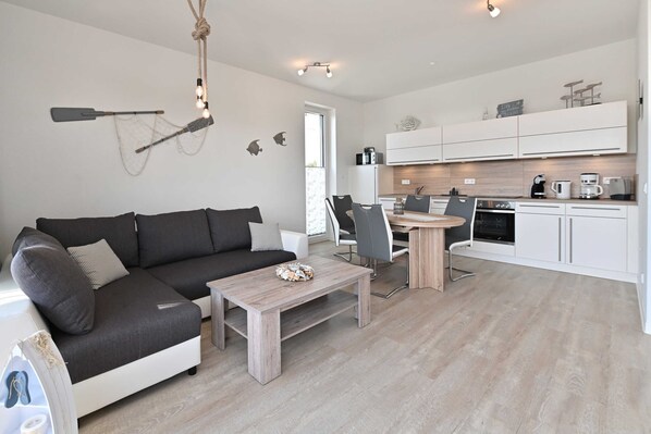 Ein modernes Wohnzimmer mit einem dunkelgrauen Ecksofa, einem Couchtisch aus Holz und einem Essbereich mit kleinem runden Tisch und Stühlen. Im Hintergrund ist eine Küche mit weißen Schränken und einer Holzarbeitsplatte zu sehen.