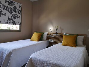 A hotel room with two beds, each with white bedding and yellow pillows. There is a bedside table with a lamp and a vase with flowers. A wall clock is visible on the table.