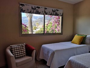 A room with two beds, a chair, and a window with a view of mountains and pink flowers.