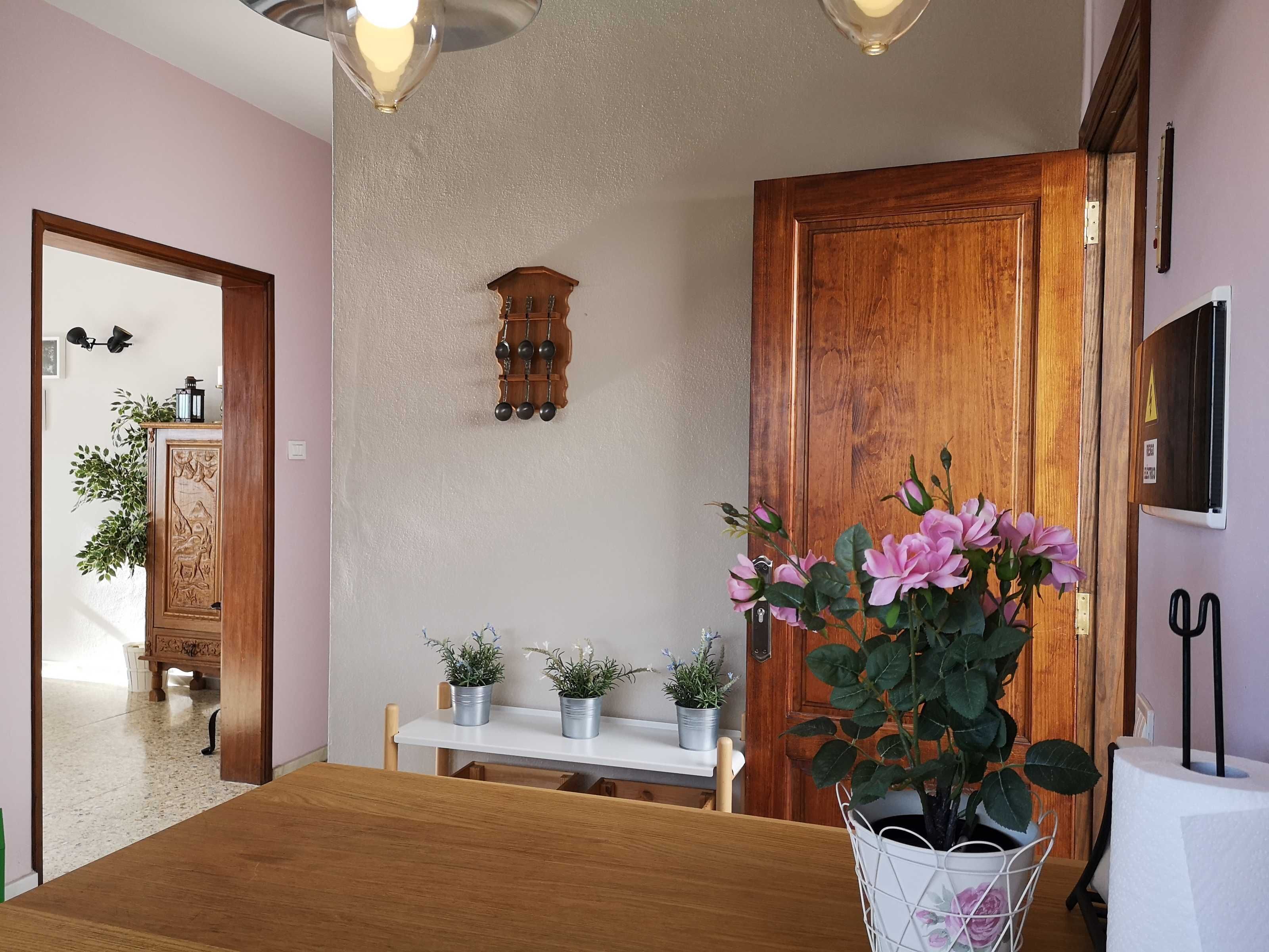 A dining area with a wooden table, chairs, potted plants, and a wall-mounted clock.