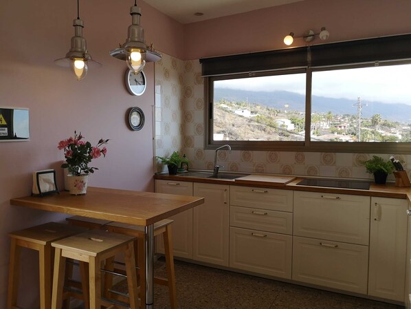 A kitchen with a wooden table, two pendant lights, a sink, and a window with a view of a town and mountains.