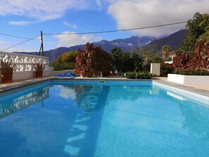 A swimming pool with clear blue water, surrounded by potted plants and trees, with mountains in the background.