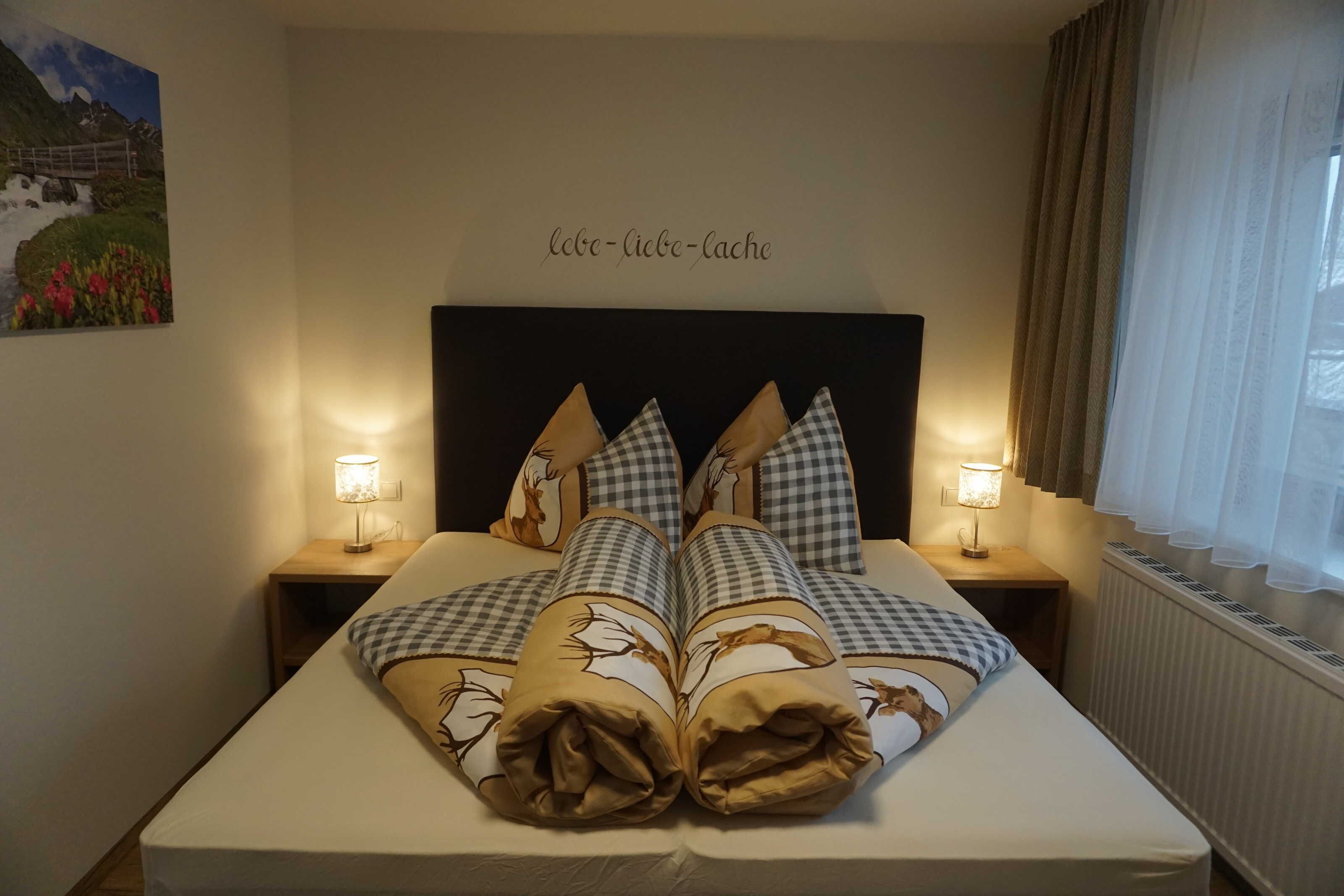 Ein ordentlich bezogenes Bett mit karierten Kissen, zwei Nachttischlampen, ein Wandbild und ein Schild mit Text.