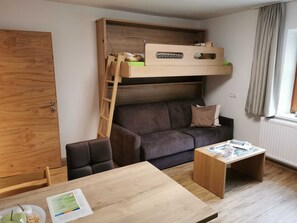 Habitación pequeña con una cama nido, un sofá, una mesa de madera y un ventanal con cortinas.