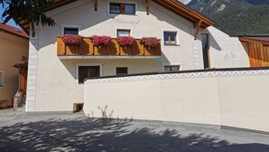 A two-story building with a balcony adorned with flowers, a mountain backdrop, and a clear sky. - Alblerhof - Apartment (2-5 people) (Pfunds)