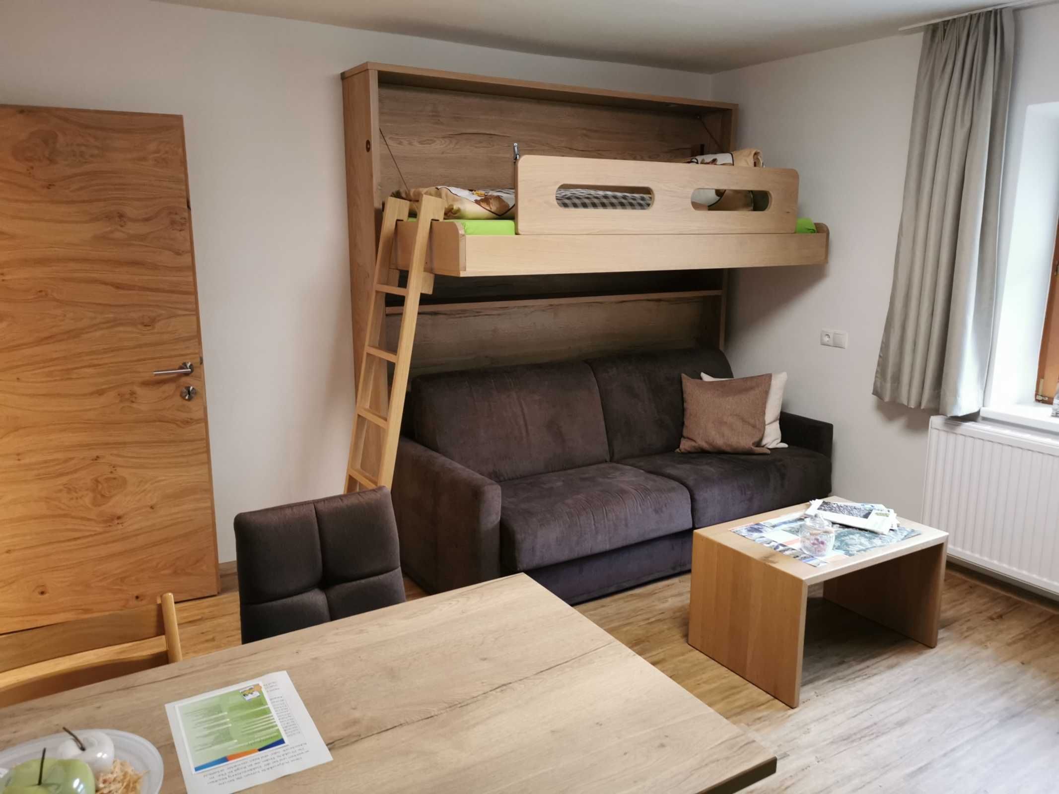 Een kleine kamer met een stapelbed, een slaapbank, een houten tafel en een raam met gordijnen.