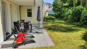 Un patio con mobiliario exterior, una mesa cubierta y un jardín con césped y arbustos.