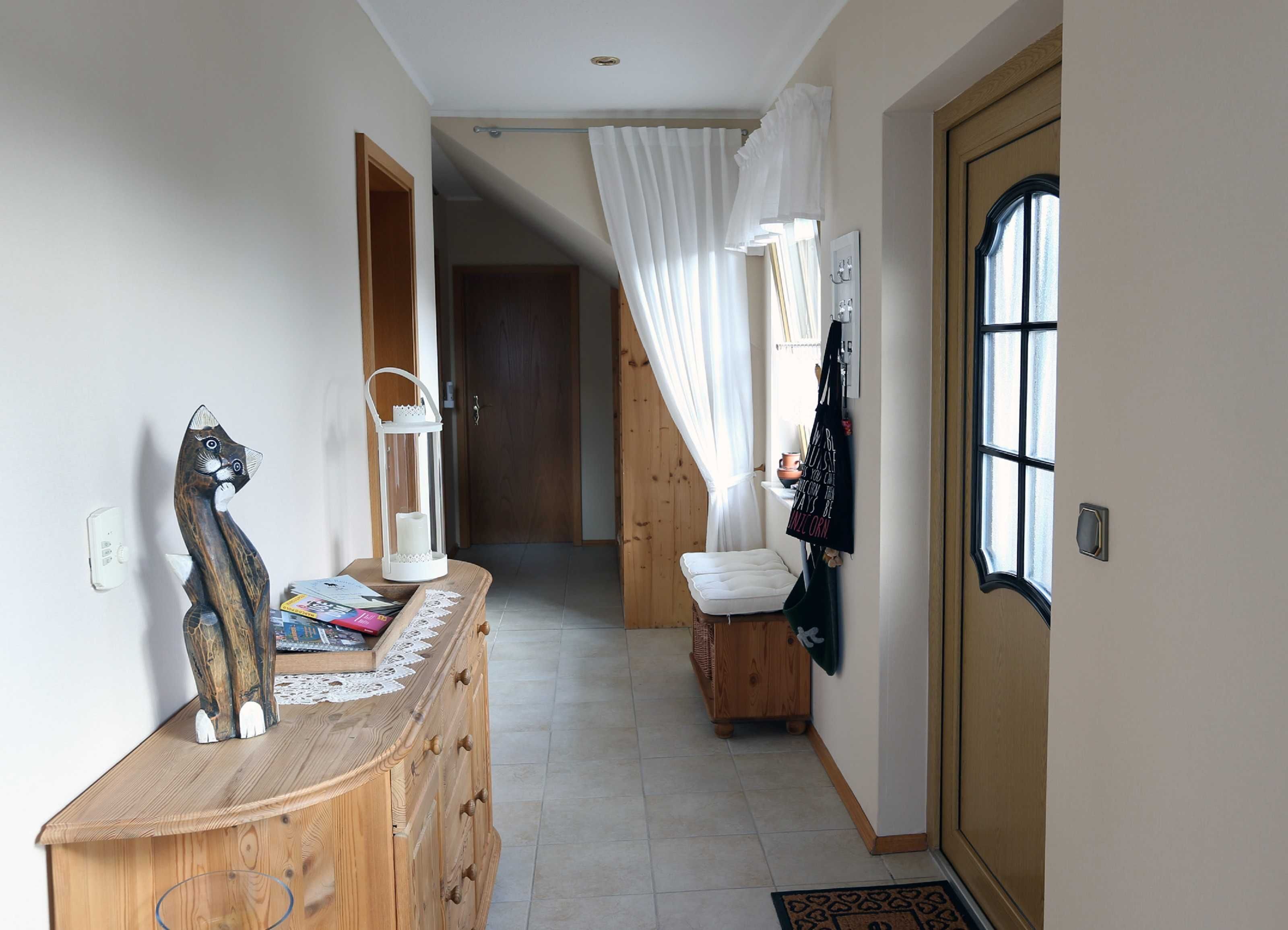 Ein Flur mit einem Holzkasten, einer weißen Katzenstatue, einem weißen Laternenlicht und einer schwarzen Tragetasche an der Wand.