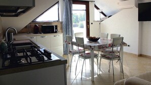 A kitchen with a gas stove, a microwave, and a sink. There is a dining table with chairs and a bowl of fruit. The room has a sloped ceiling and a window with curtains. - IL GABBIANO (IUN: P5444) (San Giovanni Suergiu)