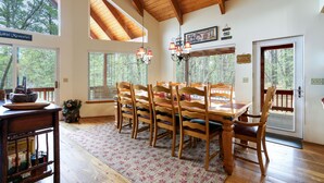 A dining area with a wooden table and chairs, large windows, and a chandelier.