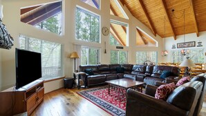A spacious living room with a large sectional sofa, wooden flooring, and a high ceiling with exposed beams.