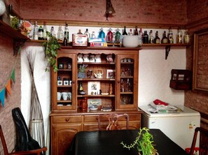 A room with a wooden cabinet containing kitchenware, a dining table with chairs, and a refrigerator.