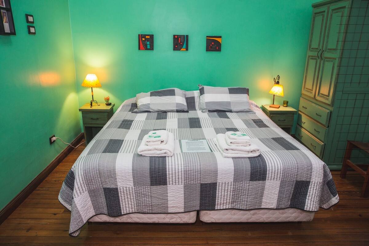 A double bed with a checkered bedspread, two bedside lamps, and a green wall with three framed pictures.
