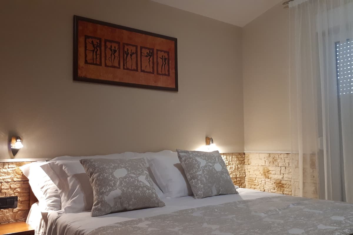 A bedroom with a stone accent wall, a bed with patterned bedding, two wall-mounted lights, and a framed artwork above the bed.