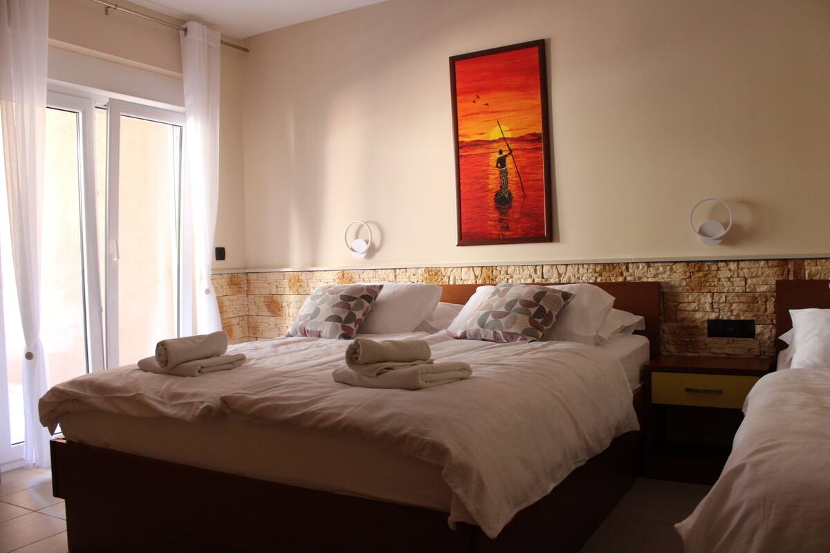 A hotel room with a large bed, two bedside tables, a painting on the wall, and a view of the outside.