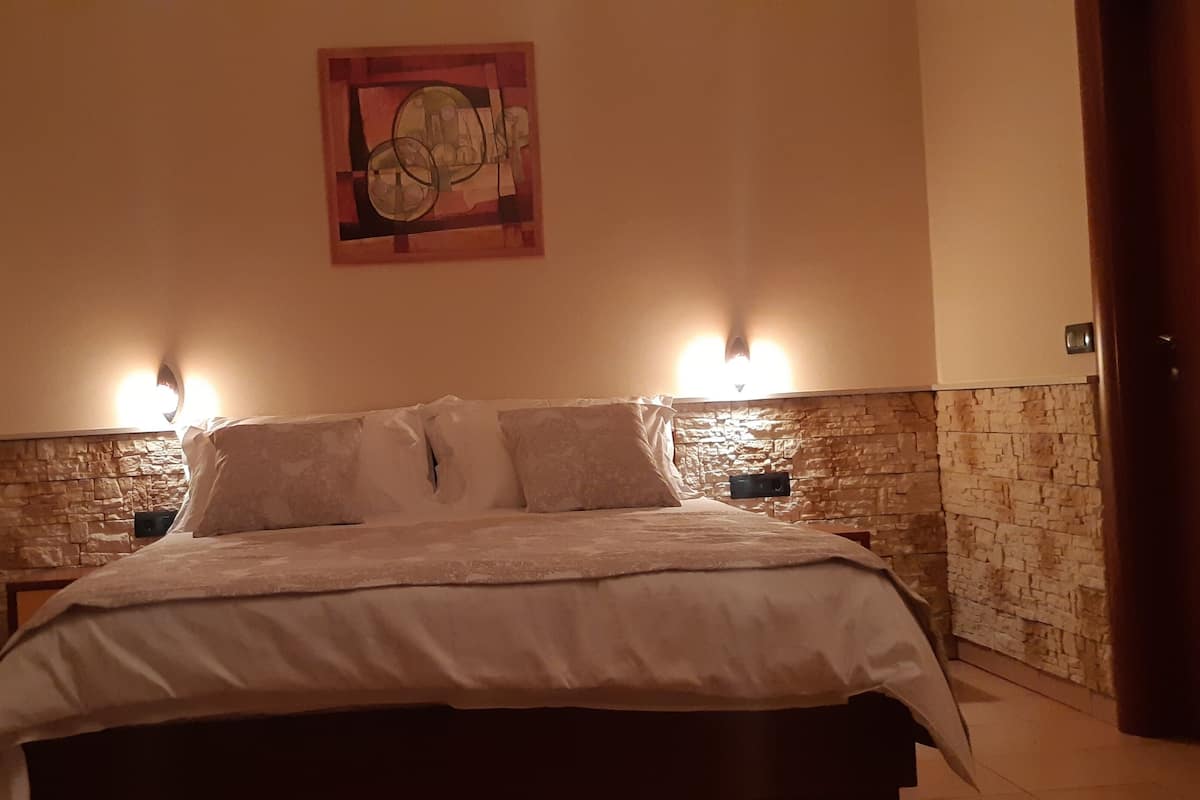 A bedroom with a stone wall, a bed with a white bedspread, two bedside lamps, and a painting on the wall.