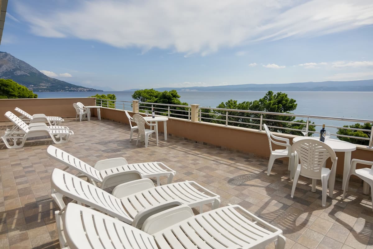 A terrace with white lounge chairs and tables, offering a view of the sea and distant mountains.