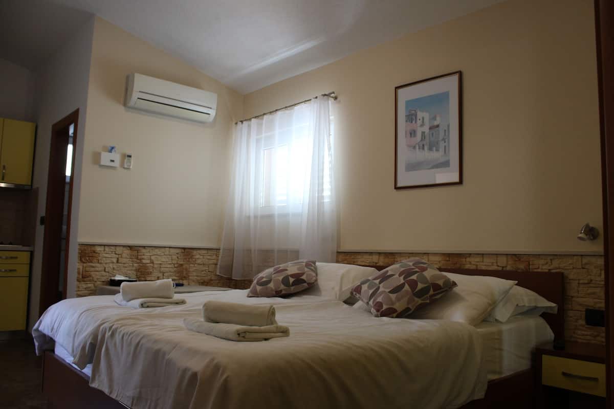 A hotel room with a bed, a nightstand, a wall-mounted air conditioner, a window with sheer curtains, a framed picture on the wall, and a stone accent wall.