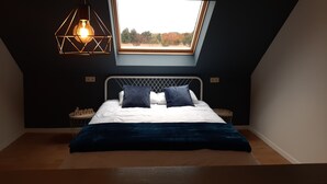 Une chambre avec un plafond en pente, un puits de lumière, un lit avec une literie blanche et deux oreillers, un plaid bleu et une suspension au design géométrique.