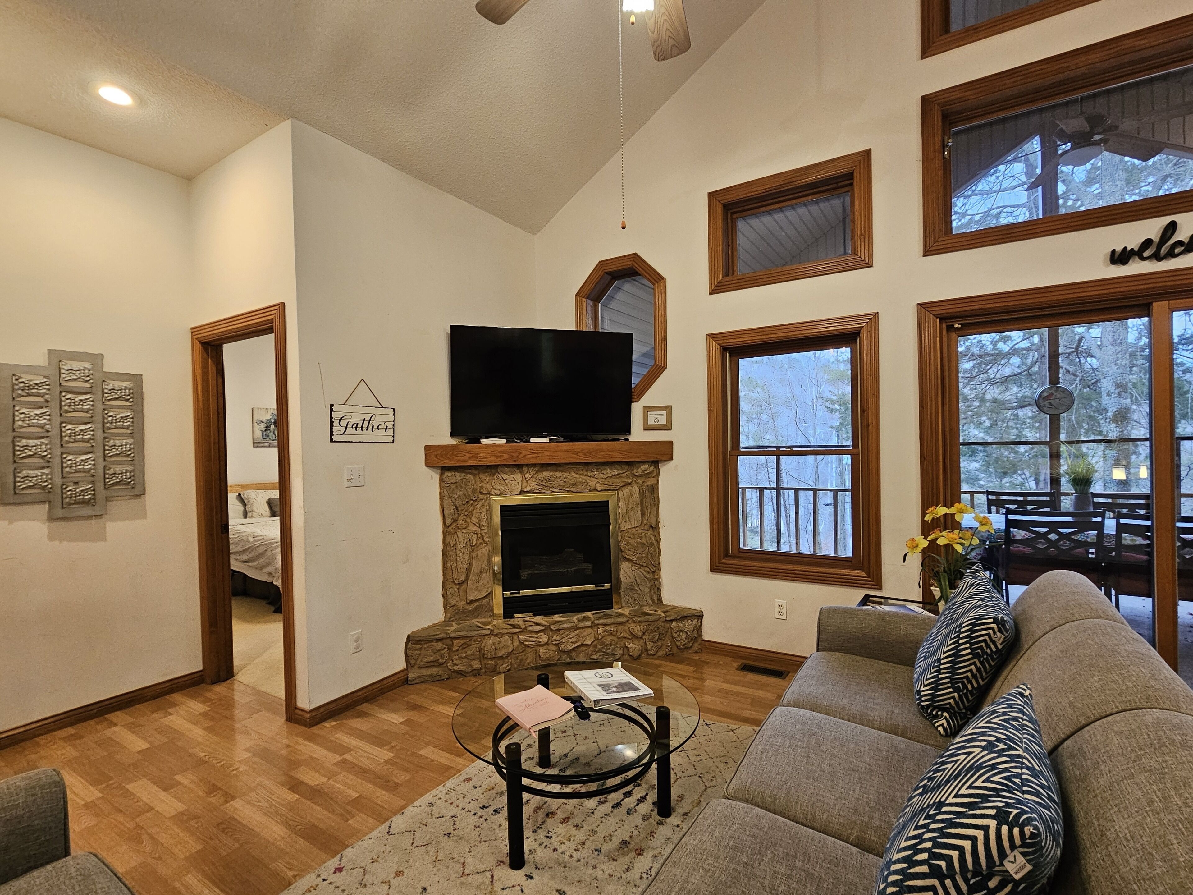 A living room with a stone fireplace, a television on top, a sofa, and a view through large windows.