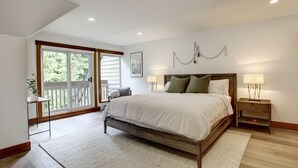 A bedroom with a large bed, two bedside tables with lamps, a view through a sliding glass door to an outdoor balcony, and a framed picture on the wall.