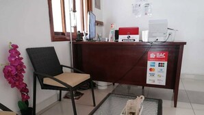 Un mostrador de recepción con una computadora, una silla, una mesita con un adorno y una planta en maceta.