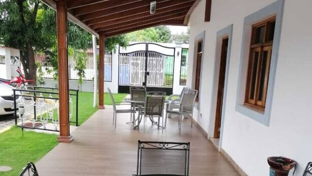 Un patio techado con una mesa metálica redonda y cuatro sillas, un pequeño adorno sobre la mesa, y una vista de un edificio blanco con una verja al fondo.