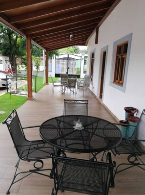 A covered patio area with a round metal table and four chairs, a small decorative item on the table, and a view of a white building with a gate in the background. - Hotel Casa Blanca (Managua)
