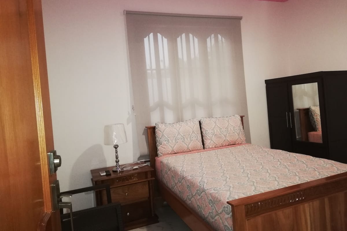 A bedroom with a bed, a nightstand, a lamp, a wardrobe, and a window with curtains.