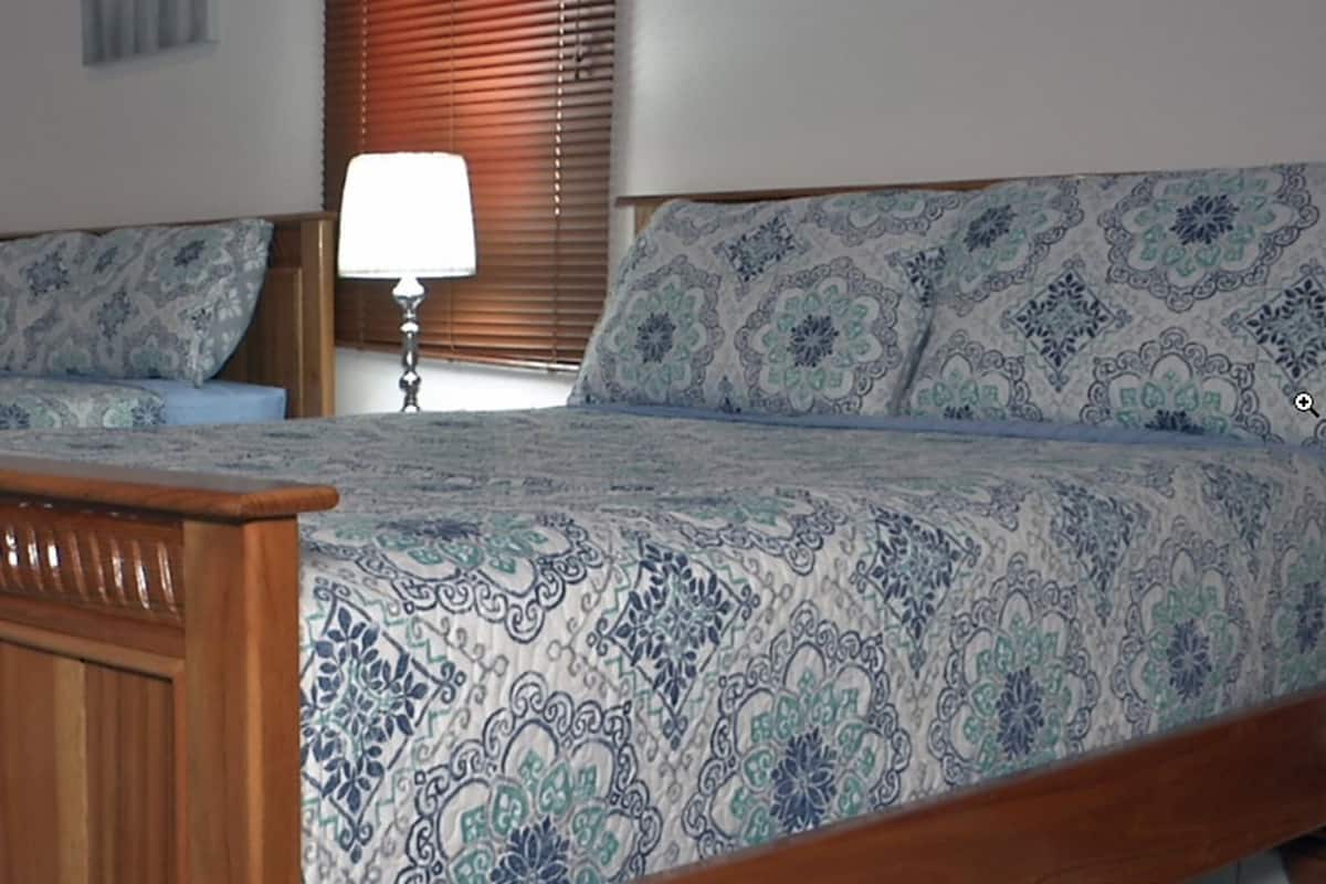 A bedroom with two wooden beds, patterned bedding, a bedside lamp, and a window with blinds.