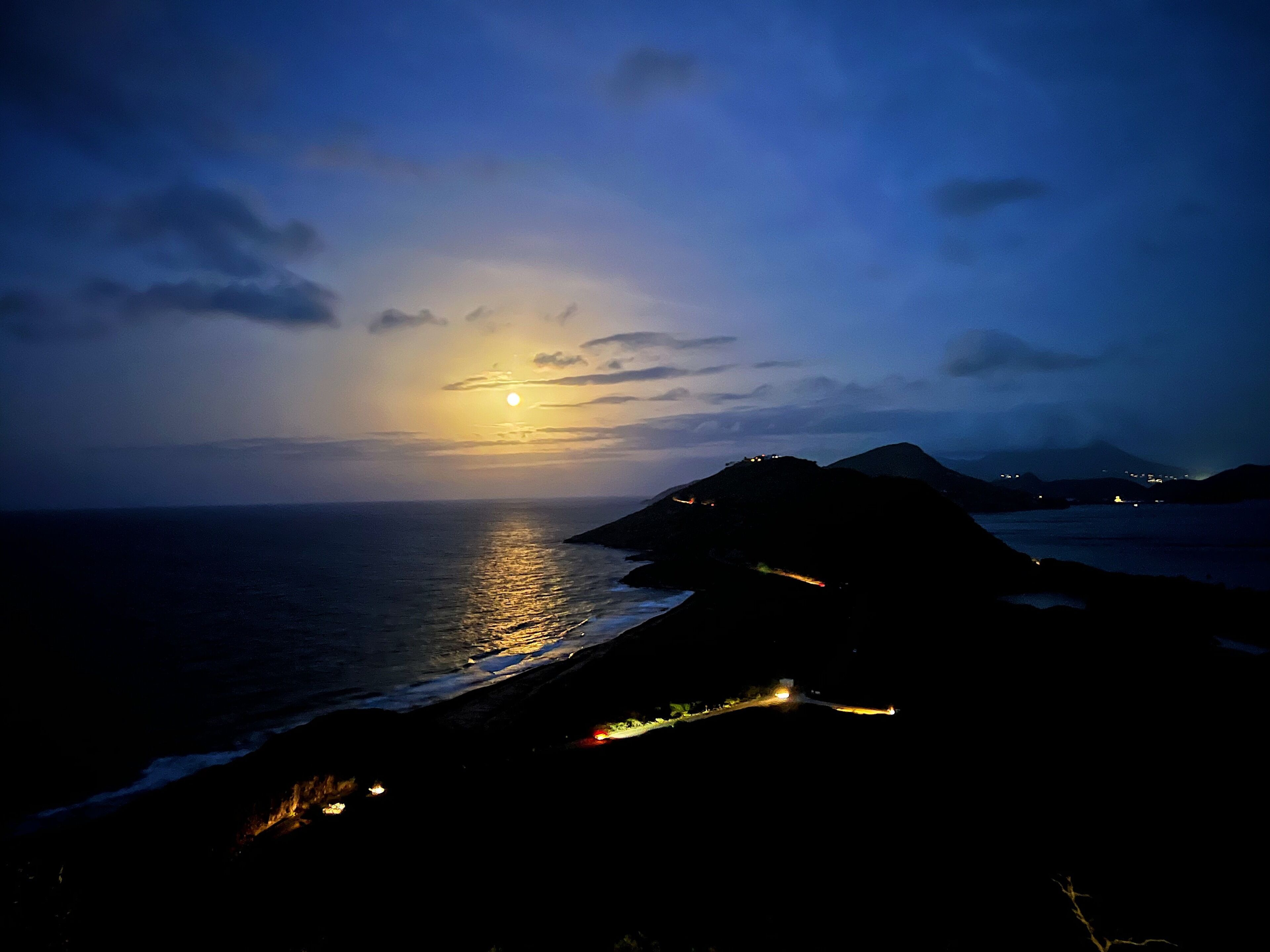 Un paysage côtier au crépuscule, avec un coucher de soleil visible sur l’océan, des bâtiments illuminés et des montagnes lointaines.