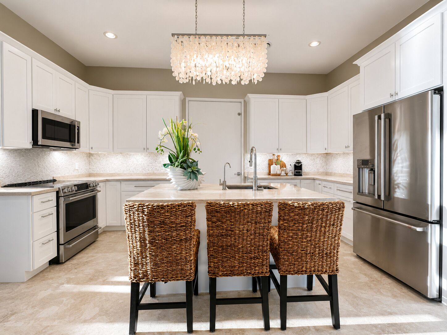 A modern kitchen with white cabinetry, stainless steel appliances, a central island with two wicker chairs, and a decorative chandelier.