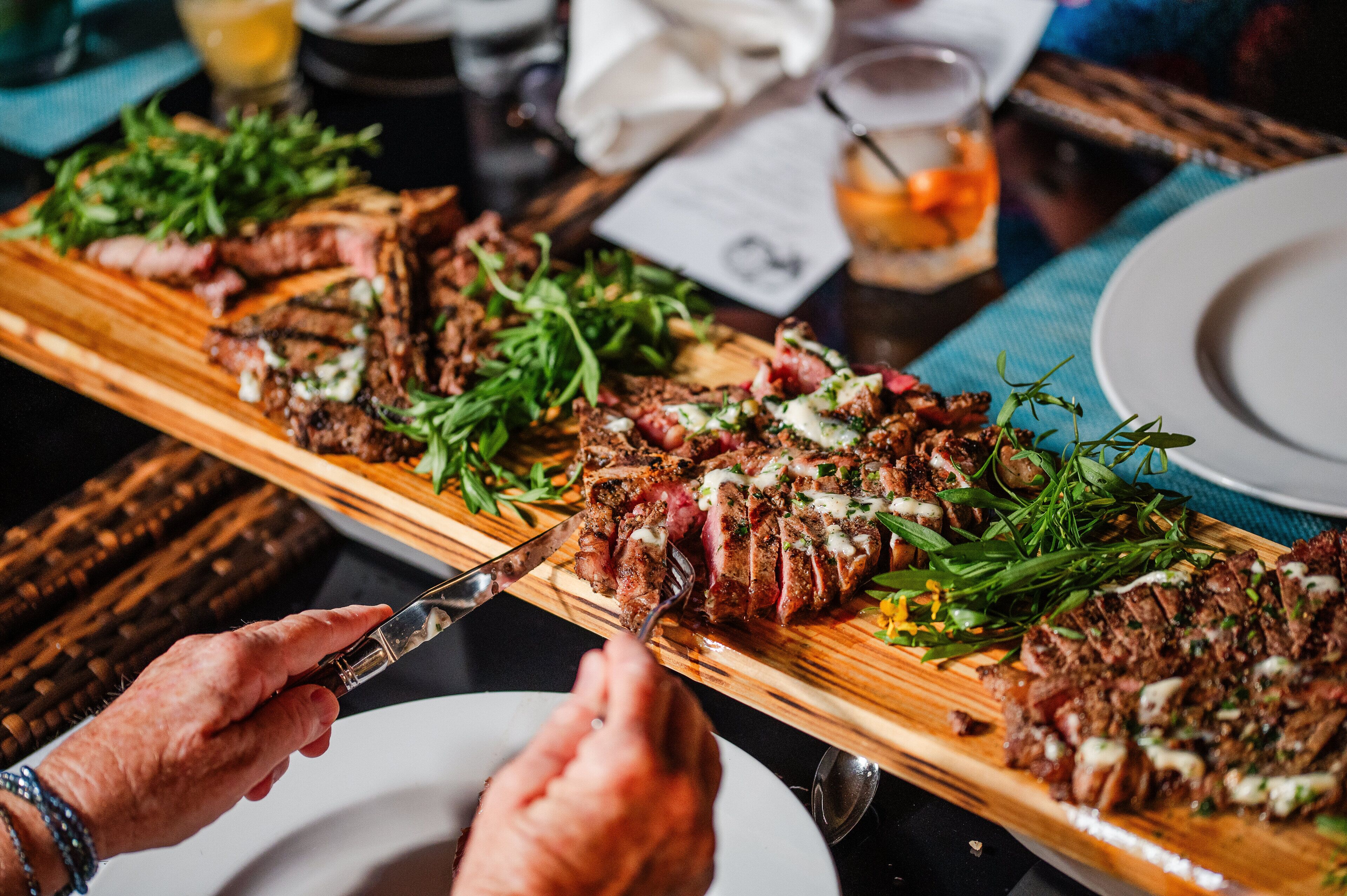 A wooden cutting board with sliced steak and greens, a knife, and a fork on a table.