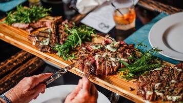 A wooden cutting board with sliced steak and greens, a knife, and a fork on a table.