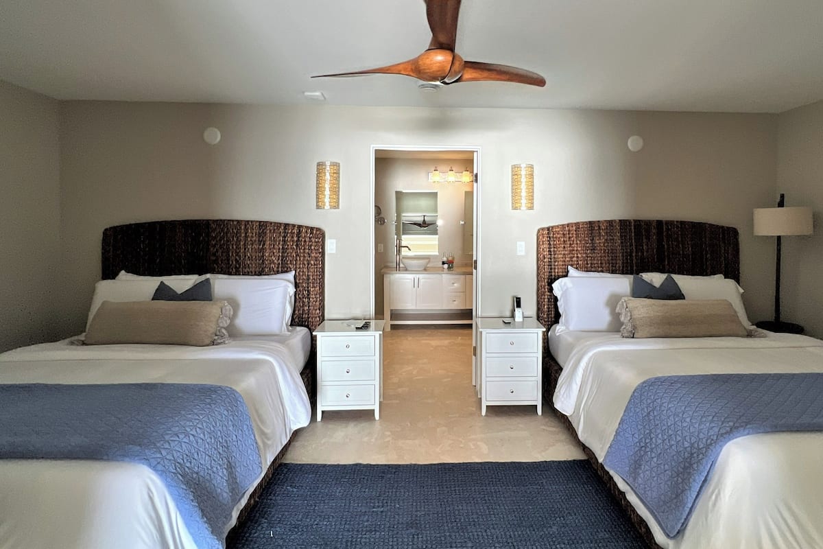 A hotel room with two beds, a ceiling fan, a nightstand, a lamp, and a bathroom visible through an open door.