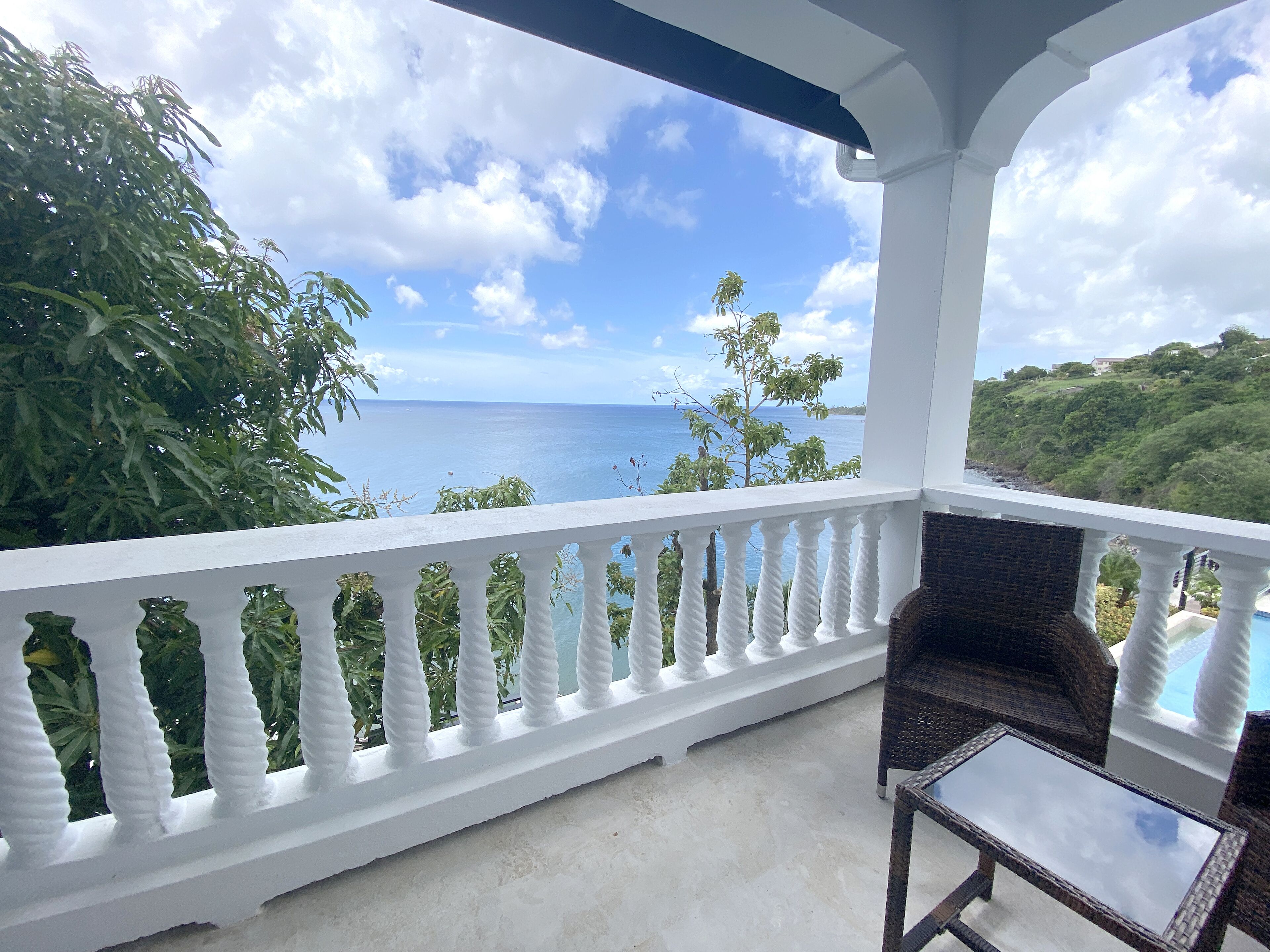 A balcony with a white balustrade, wicker furniture, and a view of the ocean and greenery.