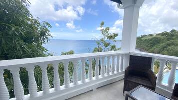 A balcony with a white balustrade, wicker furniture, and a view of the ocean and greenery.