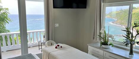 Een slaapkamer met een bed, een aan de muur bevestigde televisie, een raam met uitzicht op zee en een balkon met een stoel.