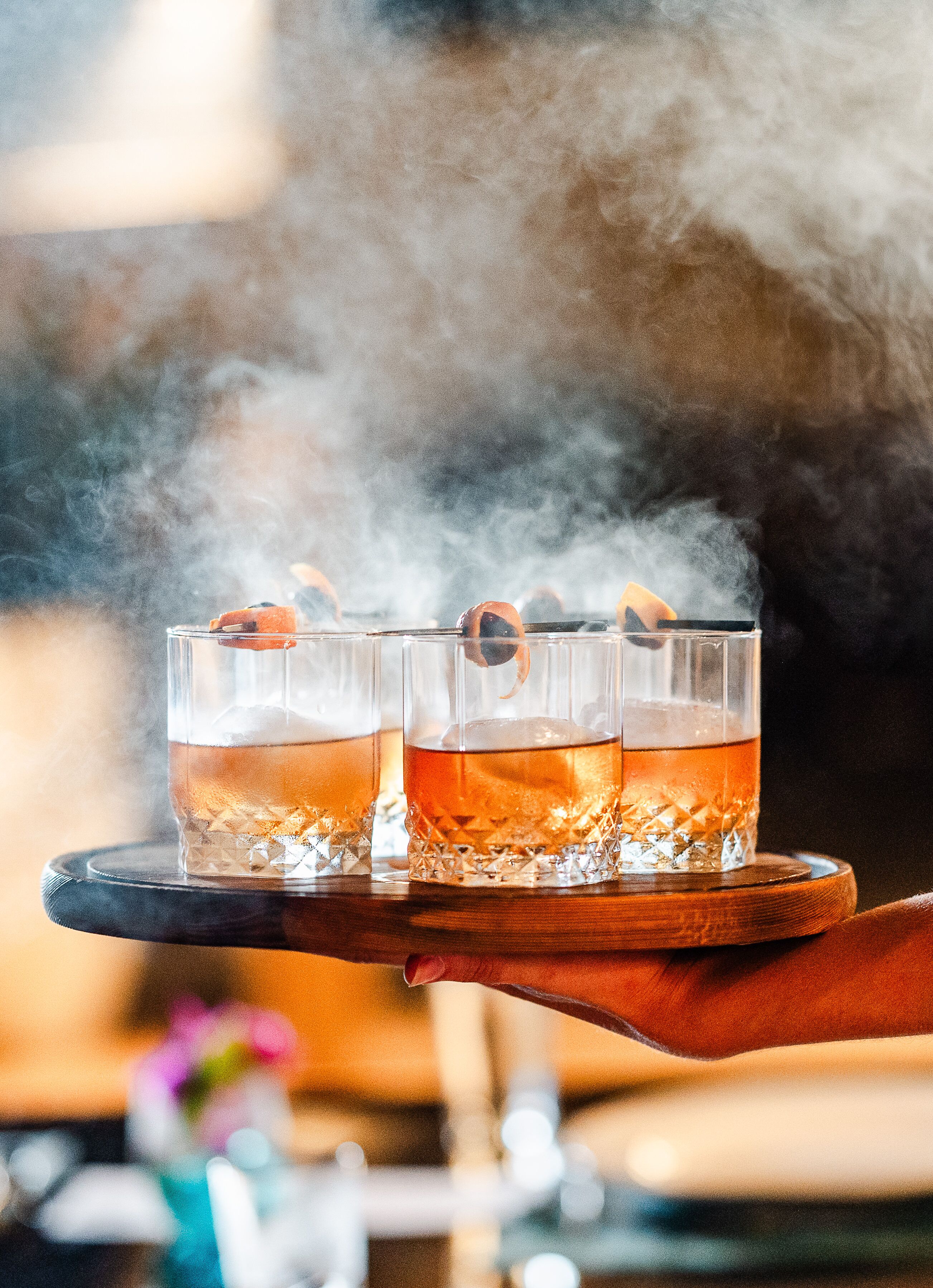 Tres vasos de líquido ámbar sobre una bandeja con cubitos de hielo, sostenida por una mano.