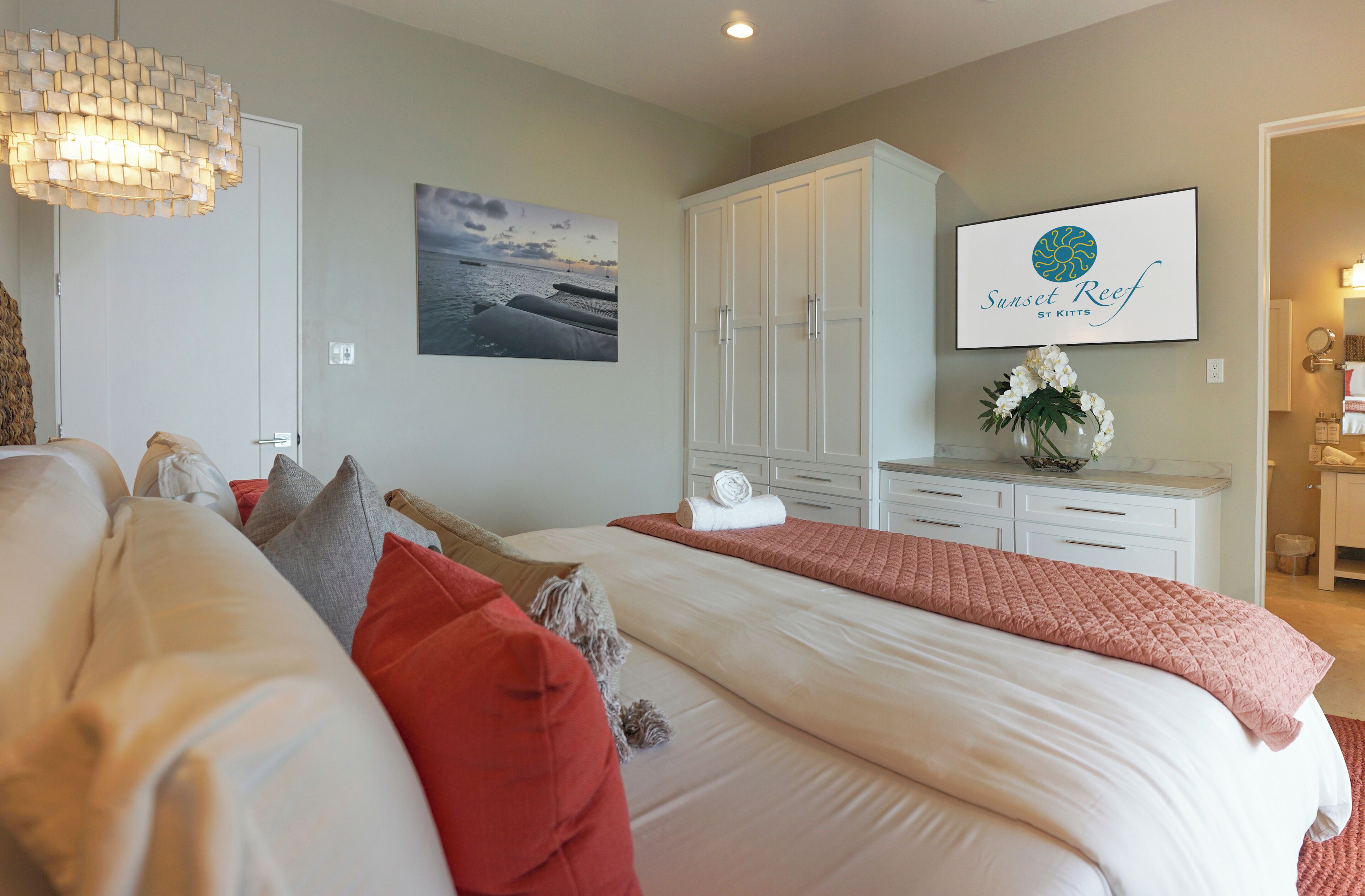 Un dormitorio con una cama grande, un armario blanco, un cuadro con una playa y un cartel que indica Sunset Reef St. Kitts.