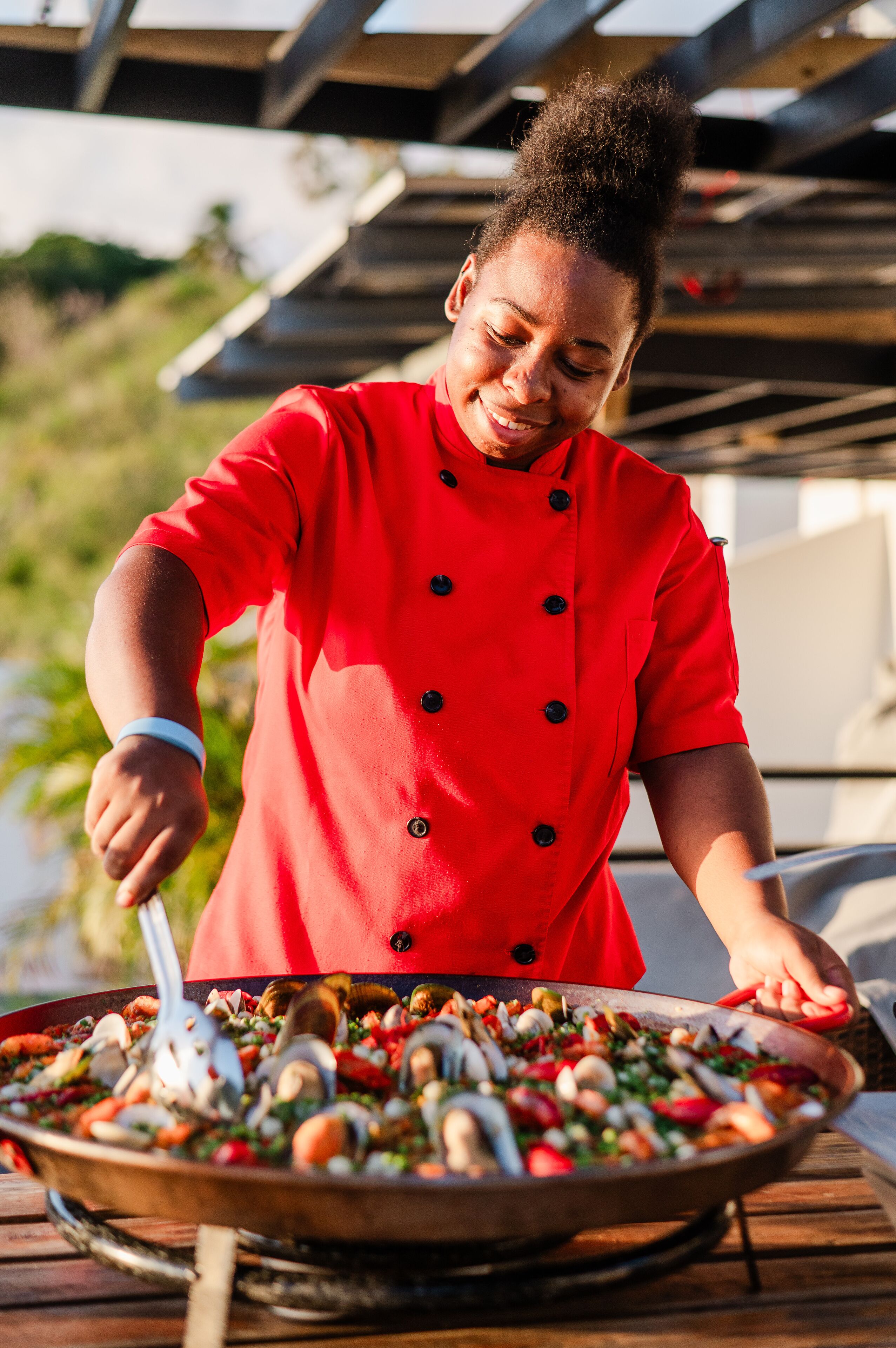 A person in a red chef's coat preparing food on a large pan outdoors.