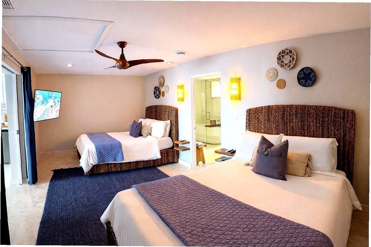 Habitación de hotel con dos camas, ventilador de techo, apliques de pared y una televisión.