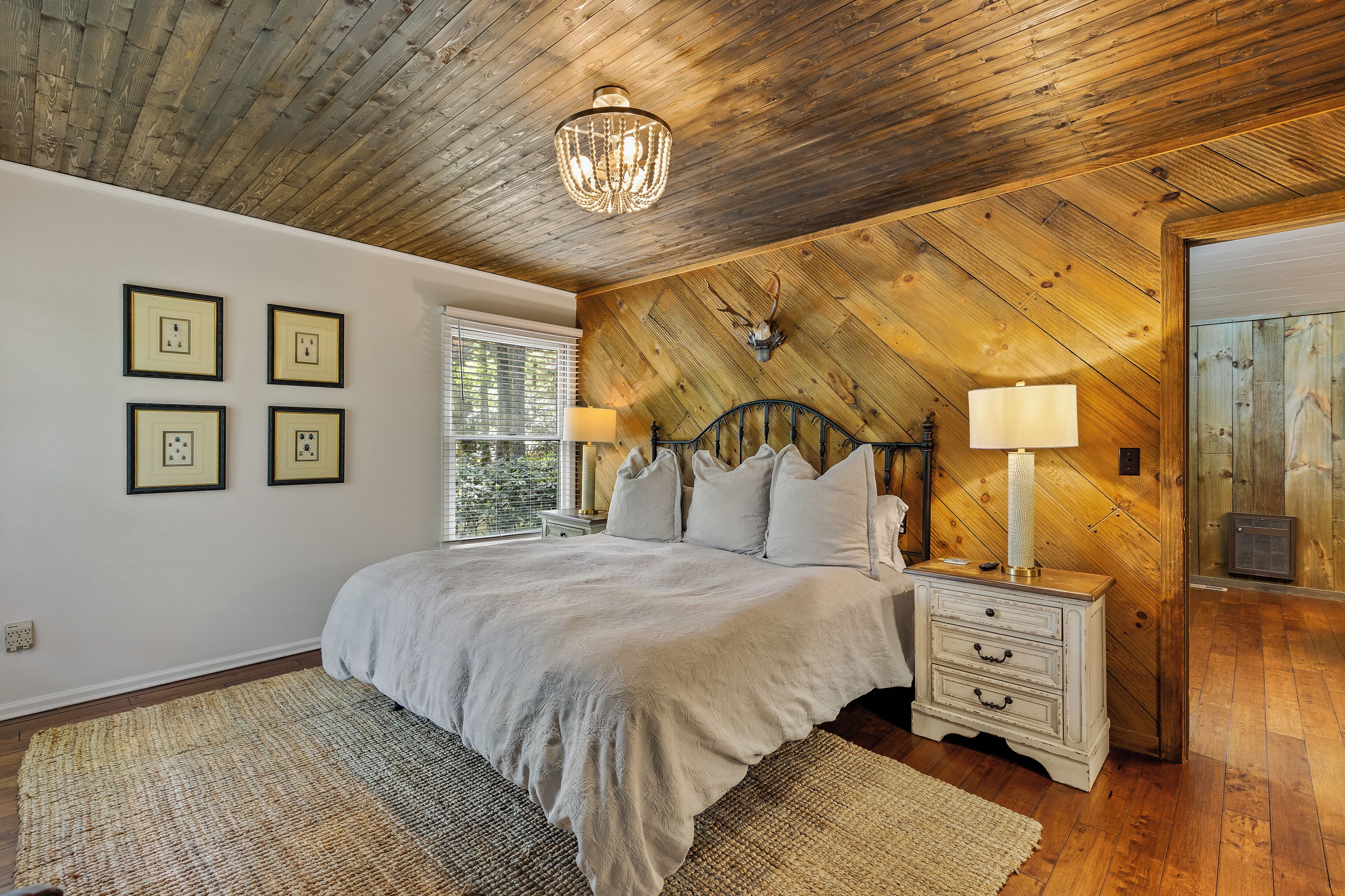 A bedroom with a wooden ceiling, a bed with a white bedspread, two bedside lamps, and a wall-mounted deer head decoration.