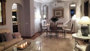 A modern living room with a glass coffee table, a sofa, a dining area with a chandelier, and a large mirror.