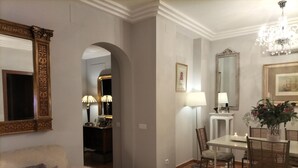 A well-lit living room with a sofa, a dining table with chairs, a chandelier, and a mirror with an ornate frame.