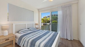 Ein Schlafzimmer mit einem Bett, Nachttischlampen, einem Weidenbeistelltisch, Blick ins Freie durch eine Schiebetür und einem Balkon mit Vorhängen.