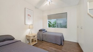 A room with two beds, a small bedside table with a lamp, a window with a view of greenery, and a ceiling fan.